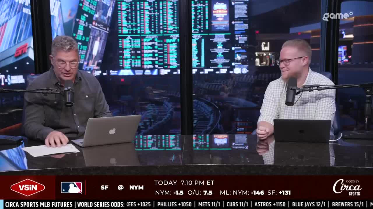 Two men sit at a desk, each with a laptop and microphone. Behind them, screens display a flurry of numbers and data, likely related to the baseball games.
