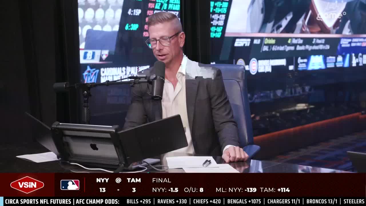 A man in a blazer and glasses speaks into a microphone, his eyes fixed on something just off-camera. The bottom of the screen displays baseball scores and betting odds, and the Game+ logo is visible in the upper right.
