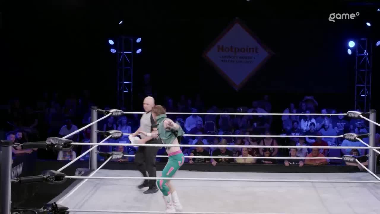 A wrestler in teal and pink is mid-motion, arm outstretched, just as the referee looks on. The crowd is visible in the background, under the bright lights of the Game+ wrestling ring.
