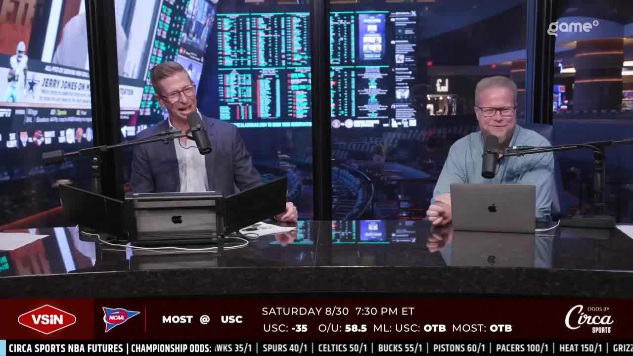 Two men sit behind a sleek, dark desk, each with a microphone and a laptop. The one on the left gestures animatedly, while the other listens intently, with the Game+ logo visible in the background.
