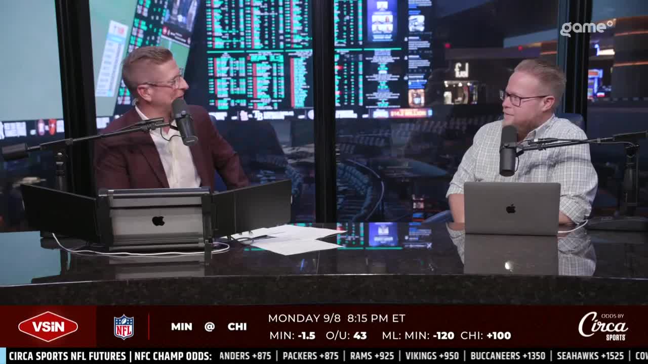 Two men in glasses sit at a dark desk, facing each other with microphones in front of them. Behind them, a screen displays what looks like sports betting information, and the Game+ logo is visible in the corner.
