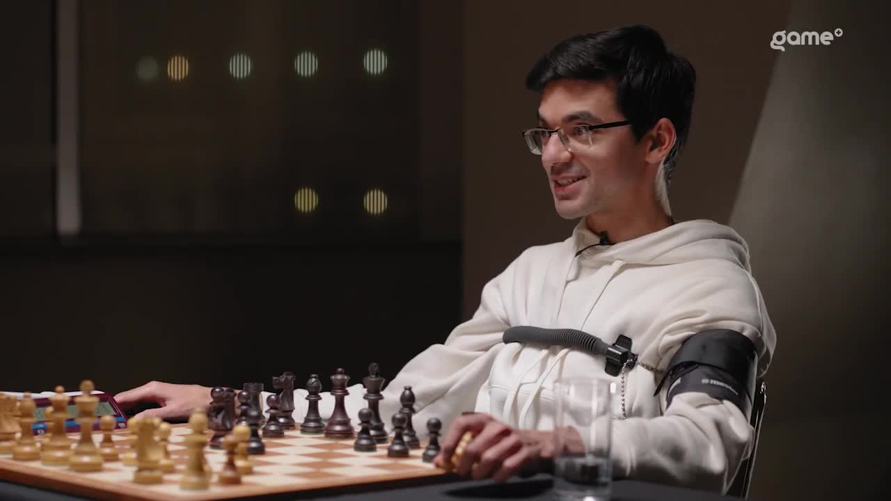 A man wearing glasses and a white hoodie is seated at a chessboard, smiling. He's got a blood pressure cuff and other sensors hooked up, likely for a Game+ broadcast.
