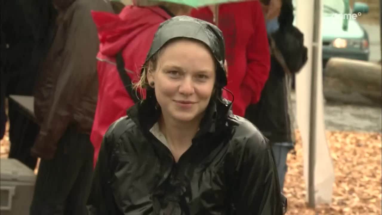 A woman in a shiny black jacket smiles at the camera, her hood pulled up against the rain. Behind her, people in red and brown coats stand under umbrellas.
