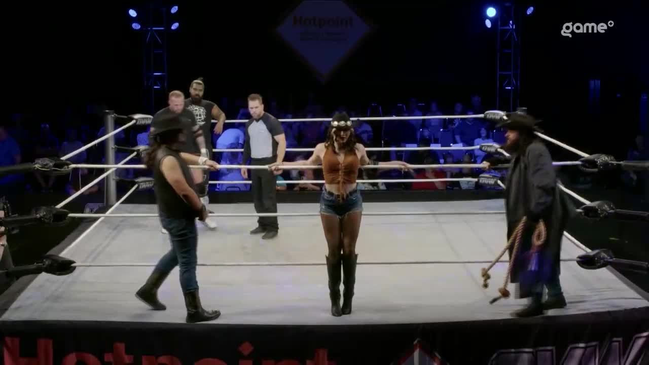 A woman in a brown vest and denim shorts stands centered in the ring, arms outstretched. Two men in cowboy hats flank her, while a referee and other wrestlers watch from the corners.
