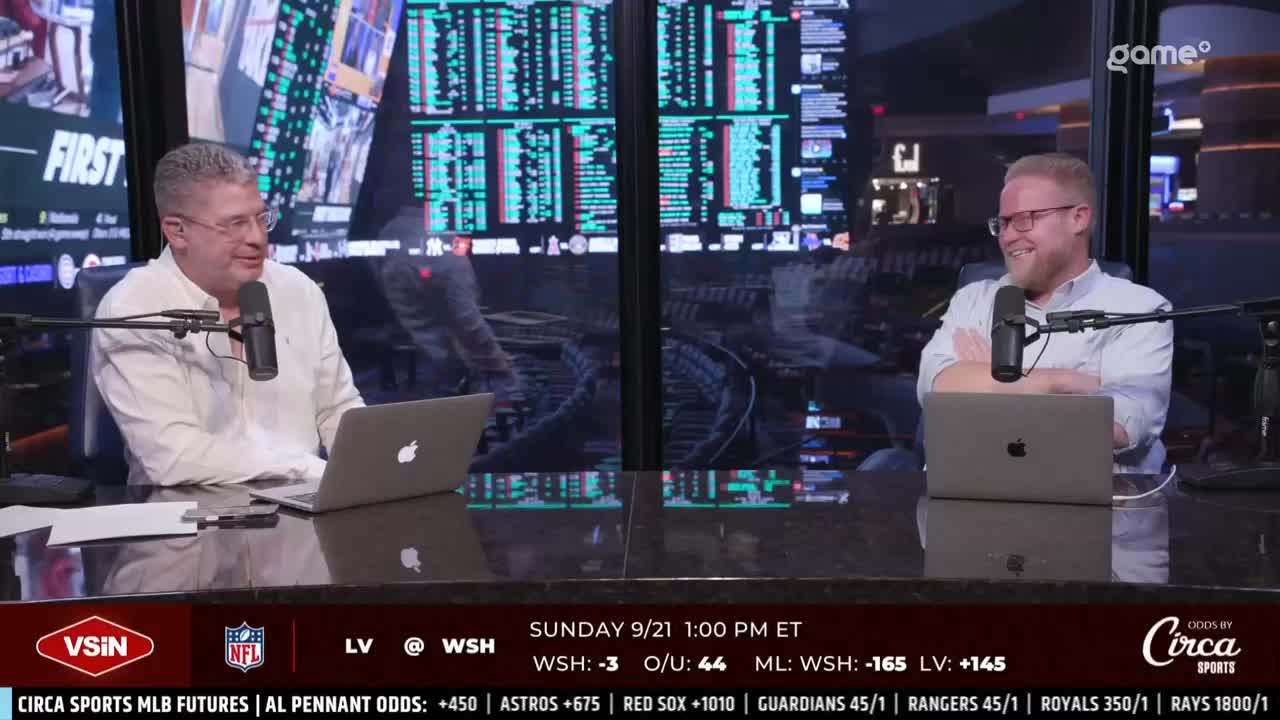 Two men, each with a laptop and microphone, are seated at a table, likely discussing sports. Behind them, a large screen displays data, and the Game+ logo is visible in the corner.
