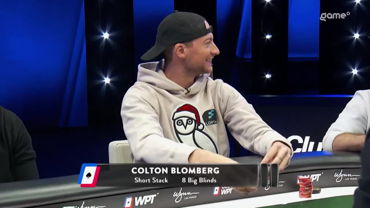 Colton Blomberg, wearing a hoodie with an owl, sits at a poker table on the Game+ set. He's smiling and looking to his right, while holding some chips.
