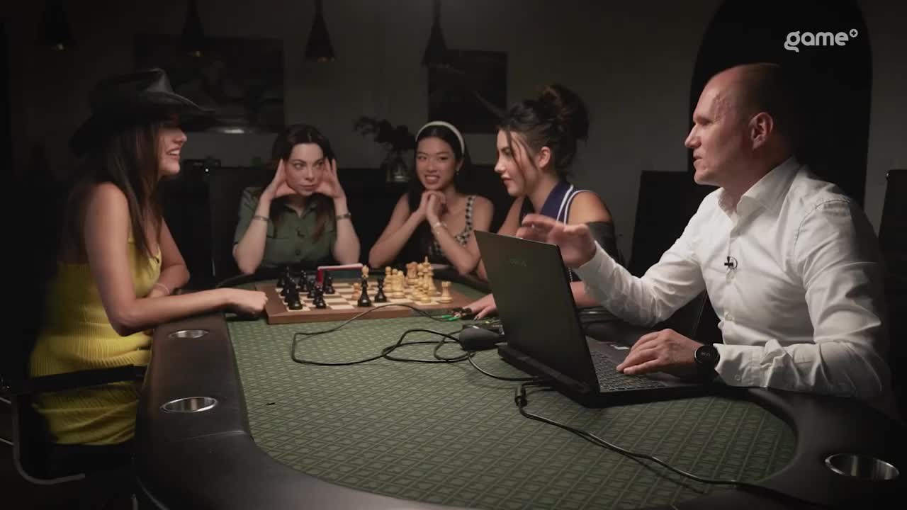 A group of people are gathered around a table, with a chessboard set up between them. The man in the white shirt gestures towards a laptop, while the others watch and listen.
