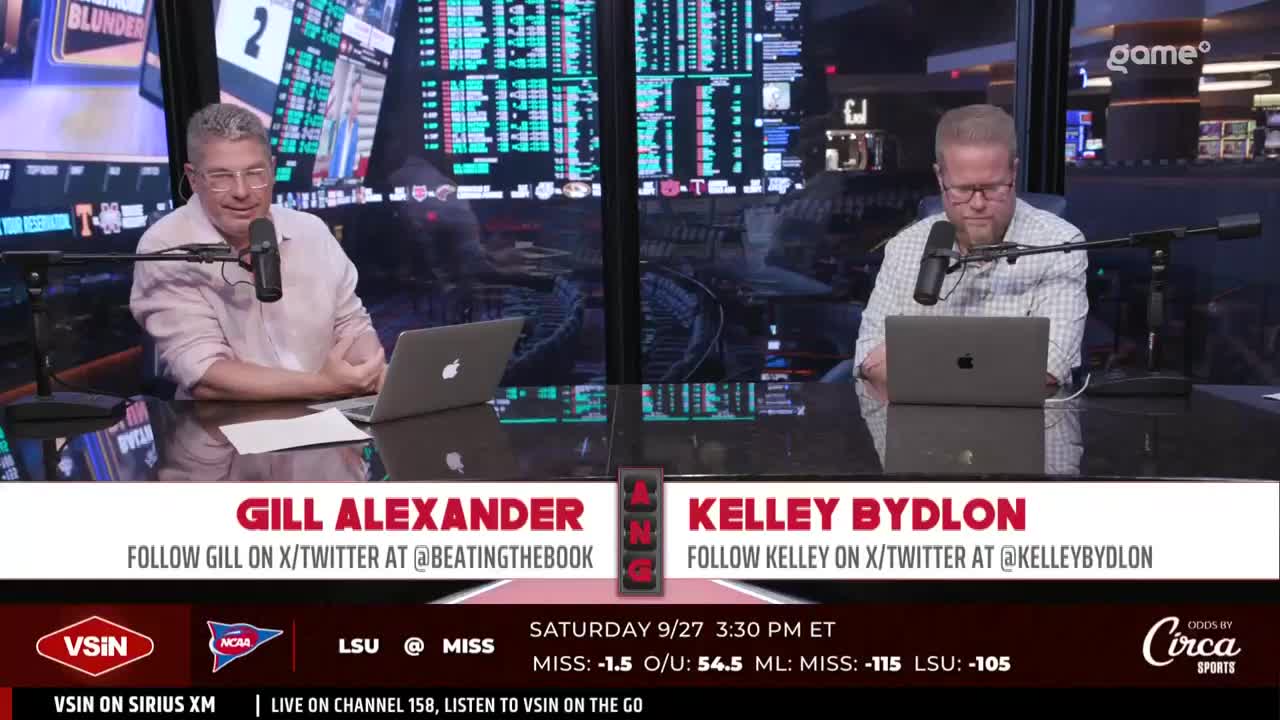 Two men sit at a desk, each with a laptop and microphone, discussing sports betting. The screen behind them displays odds and team information for an upcoming college football game.
