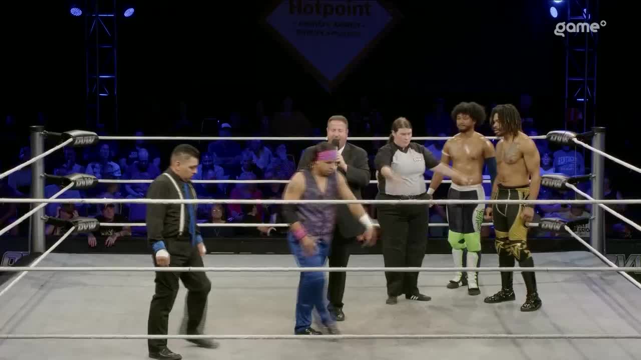 The referee gestures as two wrestlers face off, while the announcer speaks into a microphone. A third wrestler, in a vest and jeans, is being ushered away from the center of the ring.
