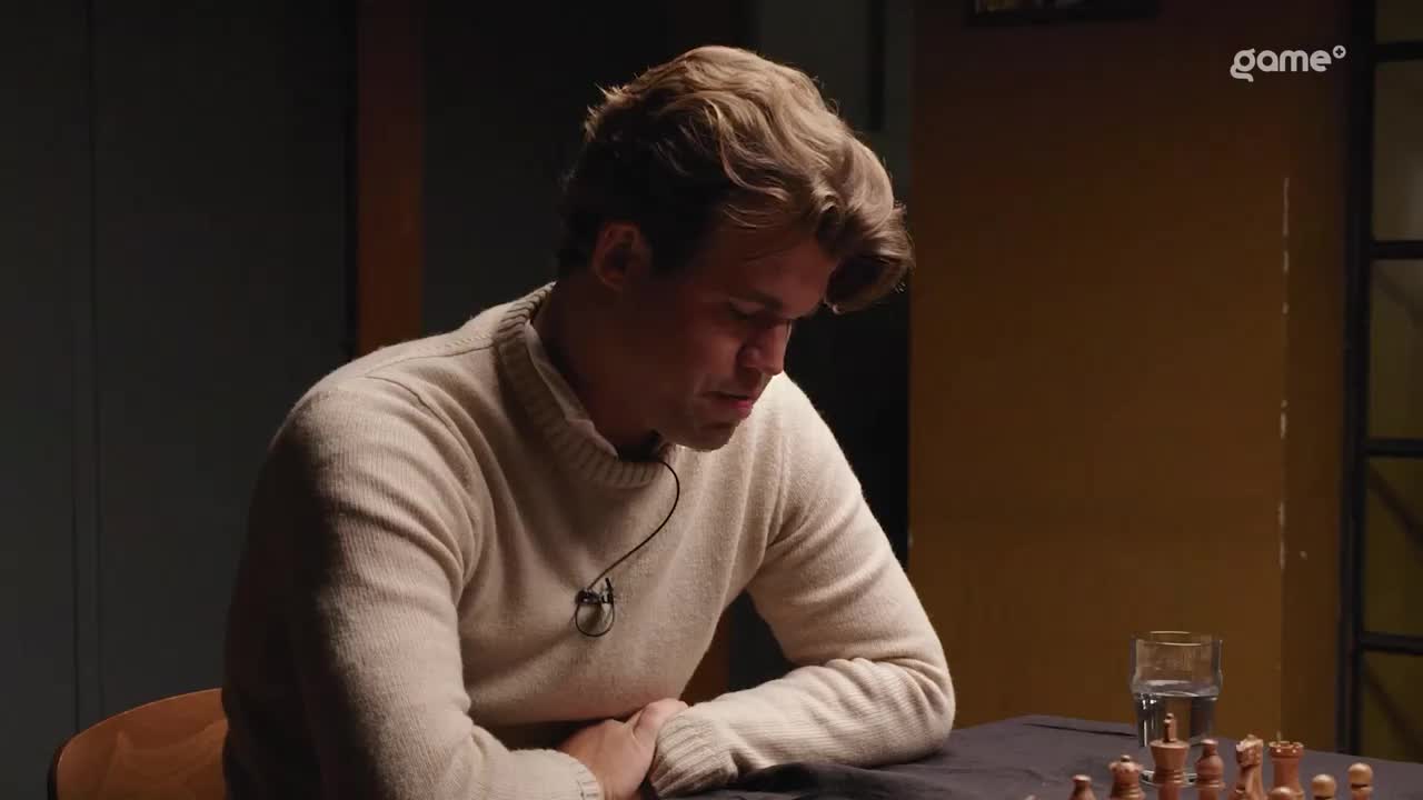 The man in the cream sweater is hunched over a chessboard, his gaze fixed on the pieces. A microphone hangs around his neck as he contemplates his next move.
