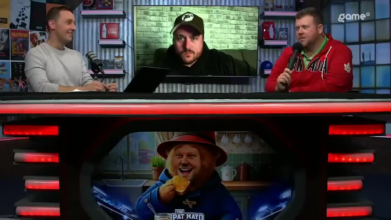 The man in the Canada hoodie is holding a microphone, speaking on a Game+ set. A screen shows another man, while a cartoonish image of a person is displayed below the desk.
