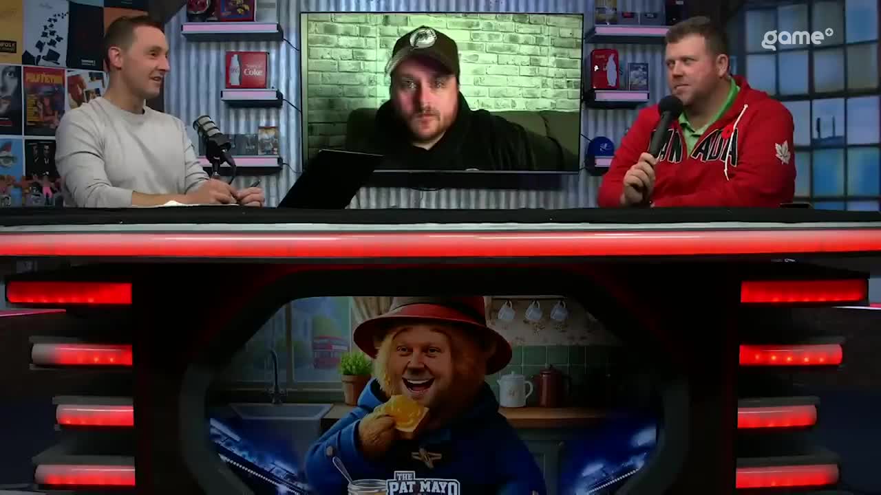 Two men sit behind a desk, one in a red Canada hoodie, the other in a grey sweater, both with microphones. A third man appears on a screen behind them, while a cartoonish figure of a bear eating a sandwich is visible on the desk's front.
