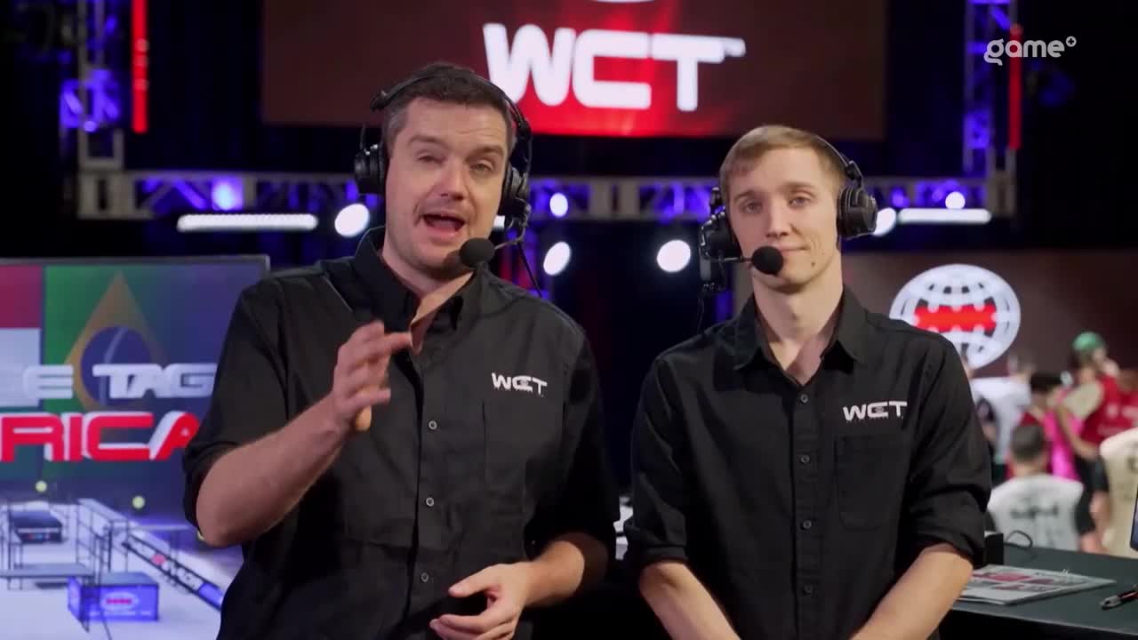Two commentators, wearing headsets and matching black shirts, are standing in front of the "WCT" logo on a large screen. The man on the left gestures with his hands as he speaks, while the other looks on.

