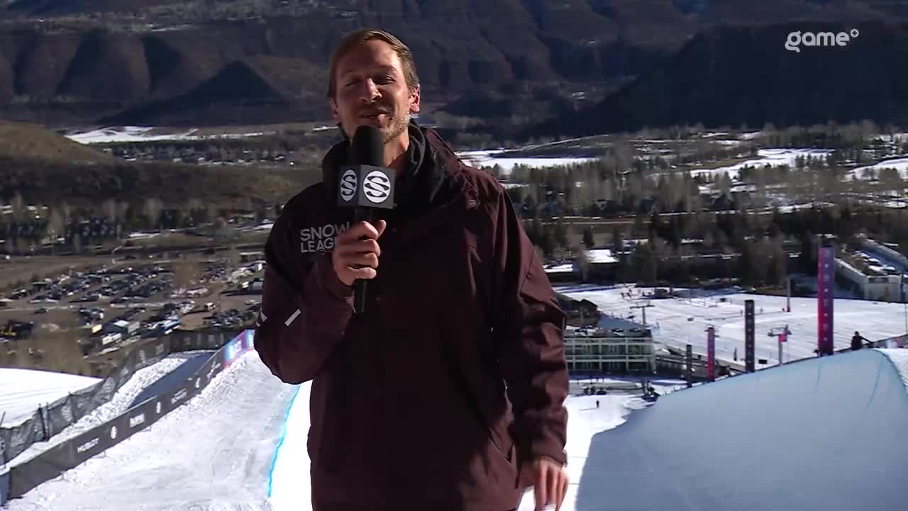 The commentator, mic in hand, is standing on the edge of the snow ramp. The backdrop is a mountain range, and the Game+ logo is visible in the corner.
