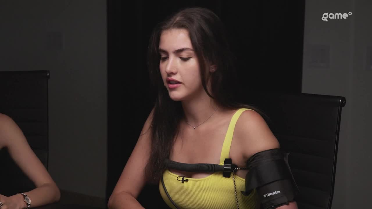 A woman in a yellow top sits in a chair, an arm cuff attached to her upper arm. She's speaking, her mouth slightly open, as if mid-sentence.
