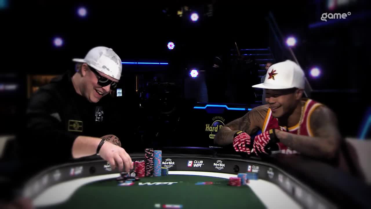 The player in the black jacket is pushing chips forward, while the other man grins, his hands resting on the table. Both are wearing hats and seem to be enjoying the game.
