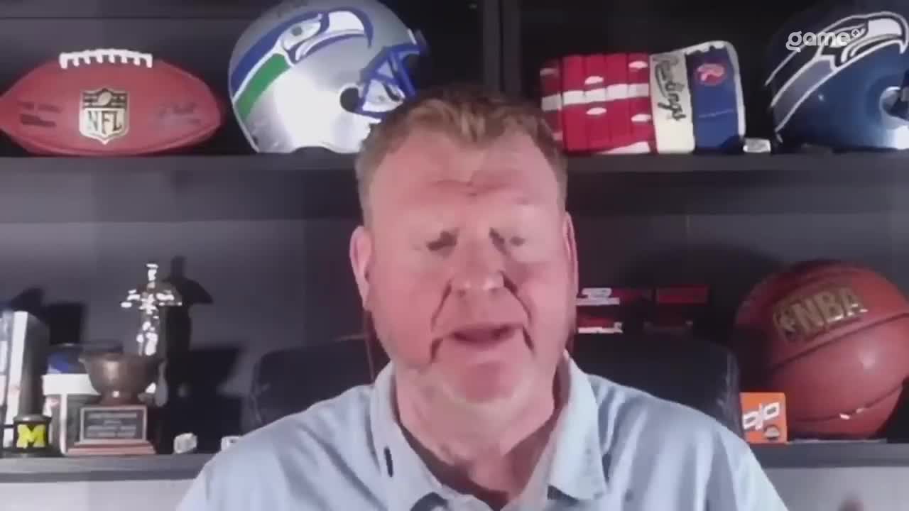 The man is speaking, his mouth open as he gestures with his hands. Behind him, shelves are lined with sports memorabilia, including helmets and a basketball.
