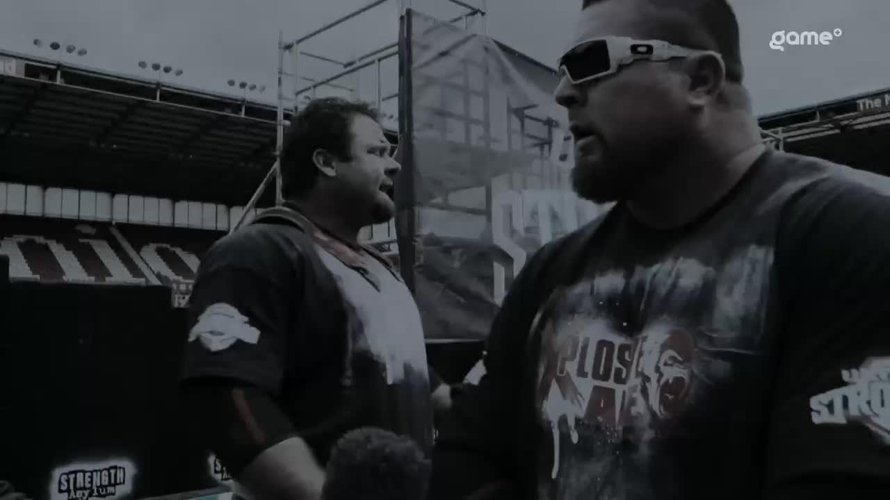 Two large men, clad in athletic gear, are engaged in a serious conversation, framed by the bleachers of a stadium. The Game+ logo peeks from the upper right corner, suggesting this is part of a strongman competition in Canada.

