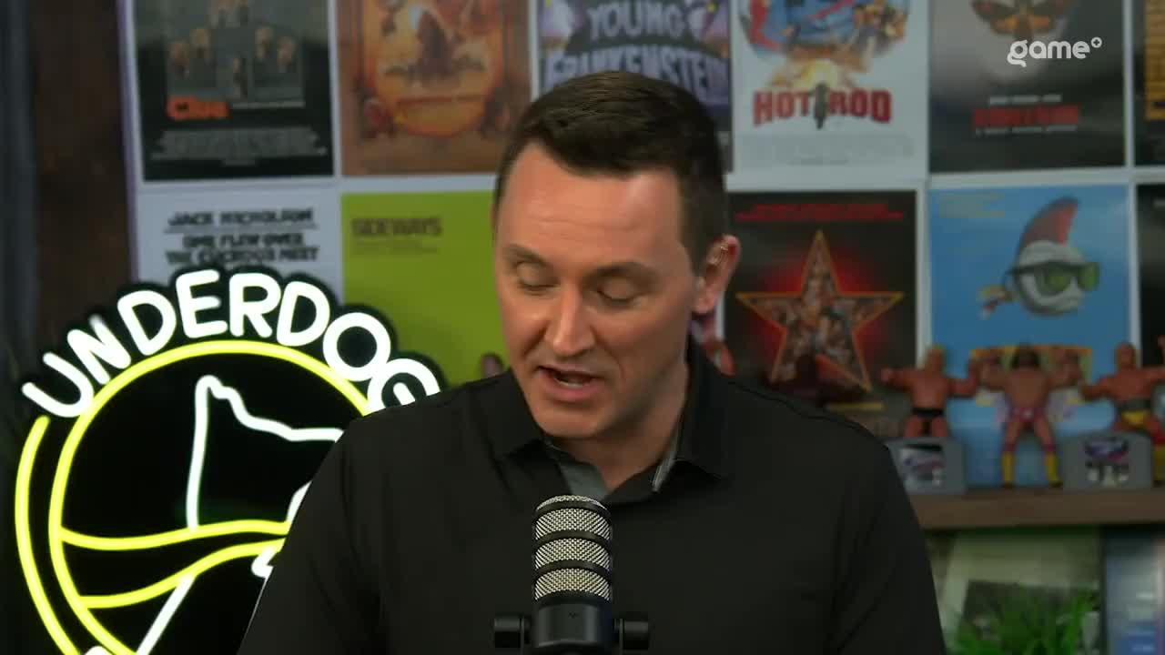 A man in a black polo shirt speaks into a microphone, with a neon "Underdog" sign behind him. The wall behind him is covered in movie posters and action figures. A man in a black polo shirt speaks into a microphone, with a neon "Underdog" sign behind him. The wall behind him is covered in movie posters and action figures.