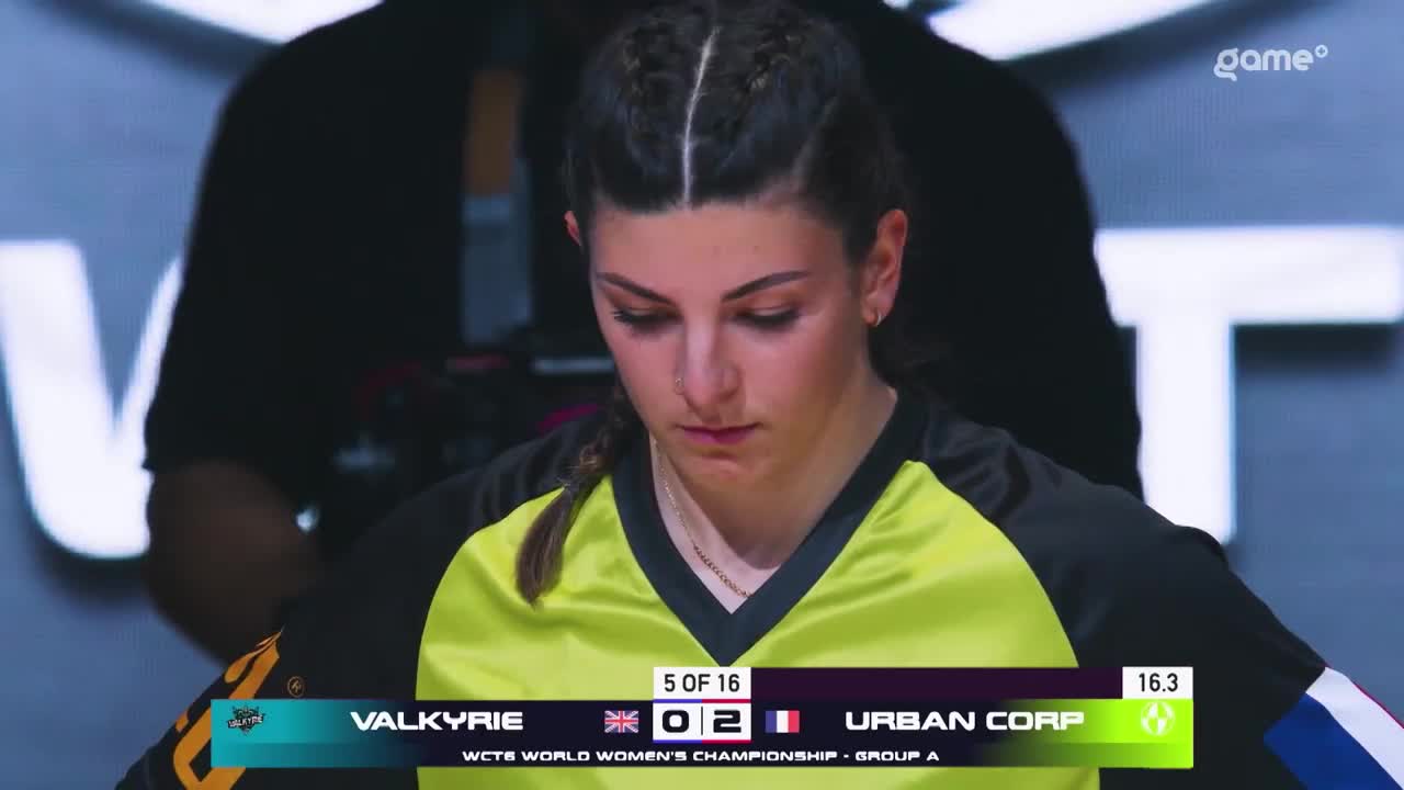The competitor, Valkyrie, stands with her head bowed, her gaze fixed on the score displayed below. The scoreboard shows "5 OF 16" and "URBAN CORP" with a score of 2, indicating a match in the WCTF World Women's Championship, Group A, broadcast on Game+. The competitor, Valkyrie, stands with her head bowed, her gaze fixed on the score displayed below. The scoreboard shows "5 OF 16" and "URBAN CORP" with a score of 2, indicating a match in the WCTF World Women's Championship, Group A, broadcast on Game+.