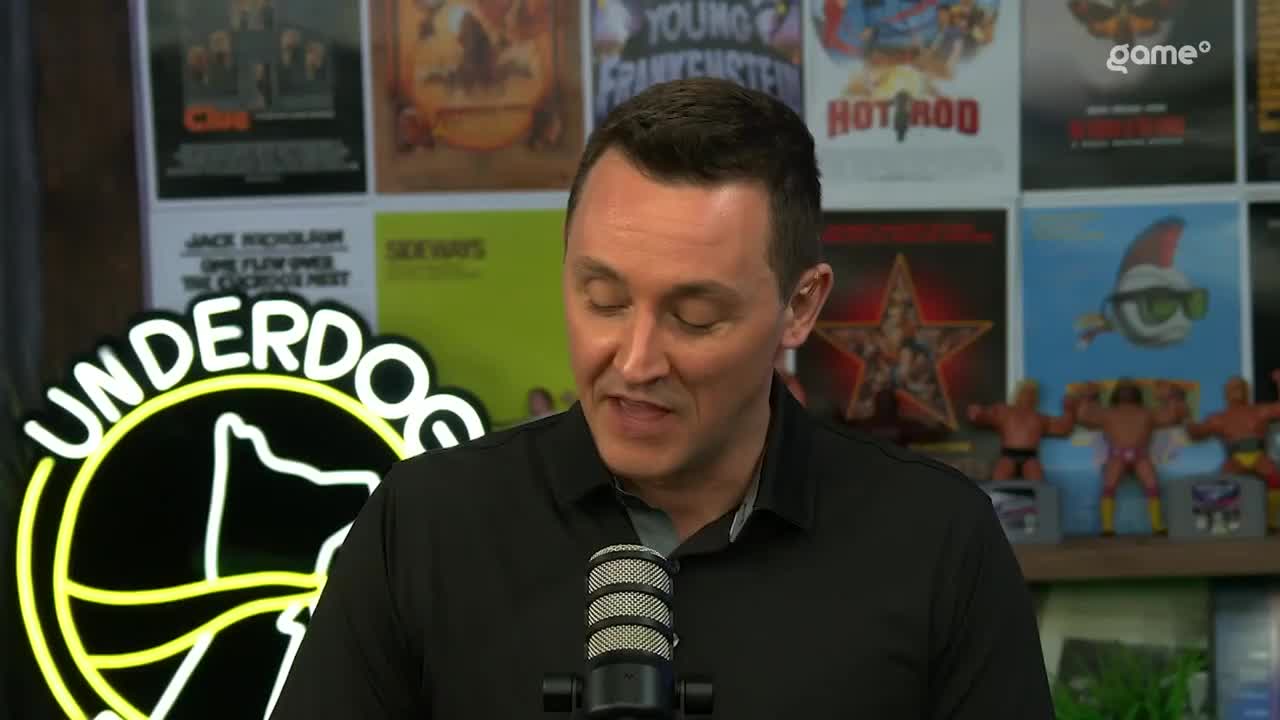 A man in a black polo shirt speaks into a microphone, with a neon "Underdog" sign behind him. The background is filled with movie posters and wrestling action figures. A man in a black polo shirt speaks into a microphone, with a neon "Underdog" sign behind him. The background is filled with movie posters and wrestling action figures.