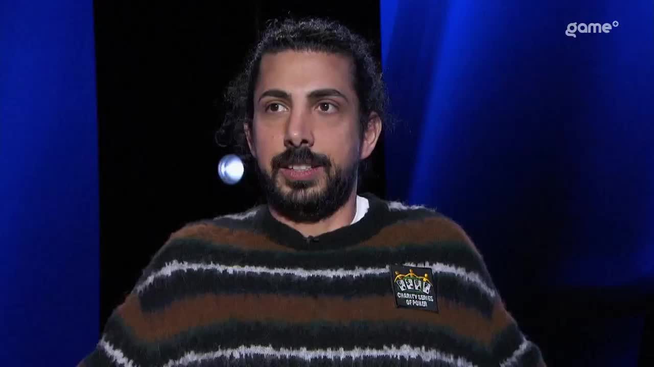 A man with dark, curly hair and a beard speaks, wearing a striped sweater. The Game+ logo is visible in the background. A man with dark, curly hair and a beard speaks, wearing a striped sweater. The Game+ logo is visible in the background.