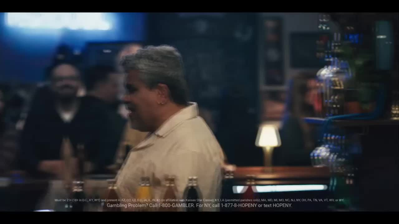 A man with graying hair and a light-colored shirt laughs at the bar. Bottles of drinks line the counter in front of him. A man with graying hair and a light-colored shirt laughs at the bar. Bottles of drinks line the counter in front of him.