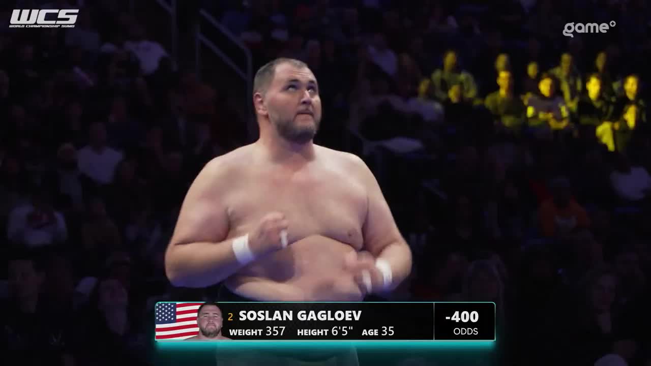 Soslan Gagloev, a wrestler from the United States, stands ready for his match, his name and stats displayed on the screen. The crowd watches intently as the Game+ broadcast continues. Soslan Gagloev, a wrestler from the United States, stands ready for his match, his name and stats displayed on the screen. The crowd watches intently as the Game+ broadcast continues.