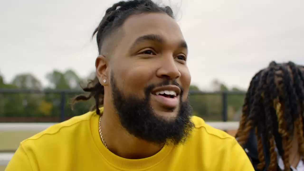 The man in the bright yellow shirt laughs, his dreadlocks swaying slightly. Behind him, another person with similar hair is visible. The man in the bright yellow shirt laughs, his dreadlocks swaying slightly. Behind him, another person with similar hair is visible.