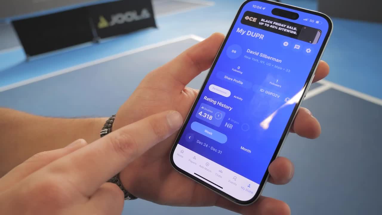 Fingers swipe across a phone screen displaying a table tennis player's rating history. A JOOLA banner is visible in the background, hinting at a tournament setting. Fingers swipe across a phone screen displaying a table tennis player's rating history. A JOOLA banner is visible in the background, hinting at a tournament setting.