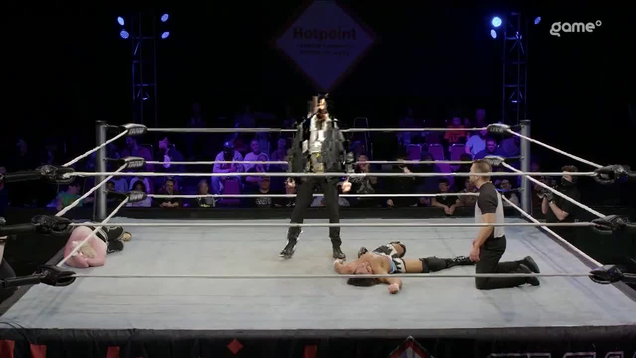 A wrestler in a black jacket stands over two fallen opponents in the ring. The referee kneels by one of the downed competitors, watching the scene unfold.