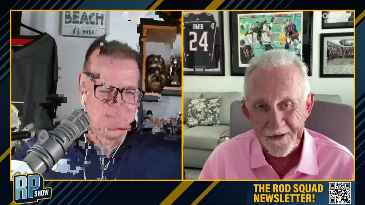 Two men are talking, one with glasses and a microphone in front of him, the other in a pink shirt. Behind the man in the pink shirt, framed hockey jerseys hang on the wall.