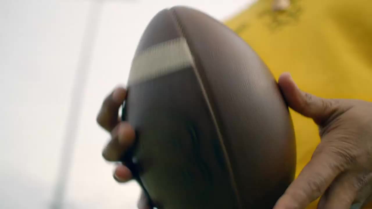A hand grips a brown football, its laces visible against the bright sky. The player's yellow jersey is a splash of color as they prepare for the next play.