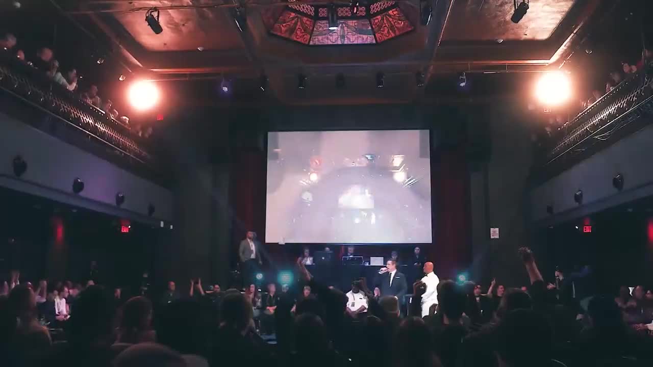 Two men in suits stand on a stage in front of a large screen, addressing a seated audience. The crowd is packed into a theater, with people also visible in the balconies above.