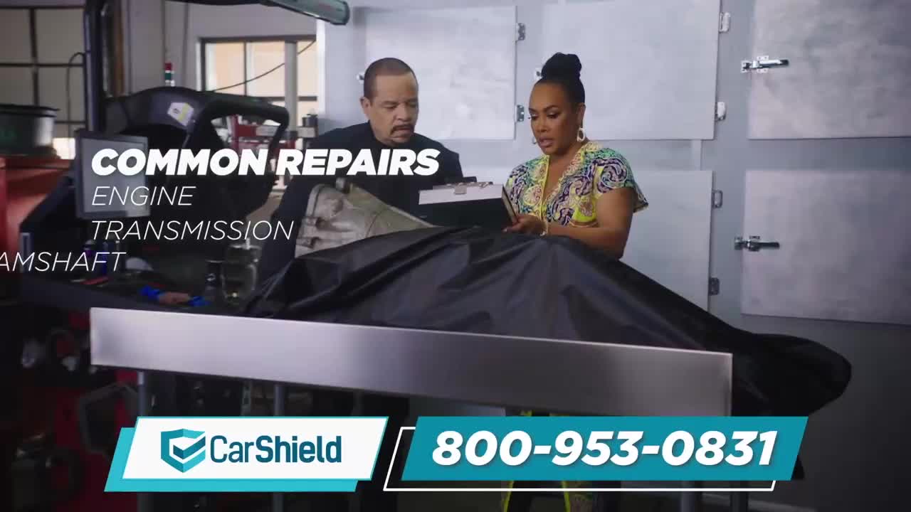 A man and woman stand over a table covered by a black tarp, looking at a clipboard. Behind them, a mechanic's bay is visible with the words "COMMON REPAIRS" and "ENGINE TRANSMISSION CAMSHAFT" displayed.