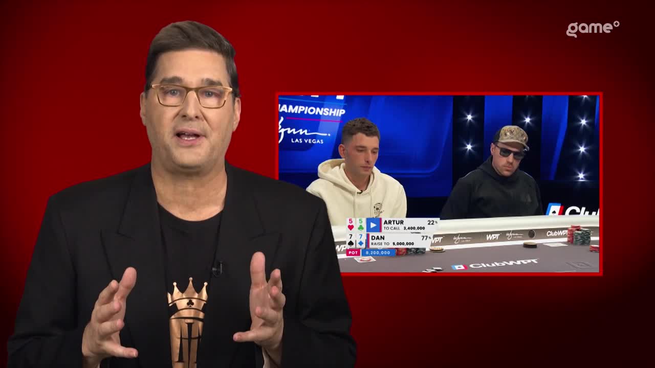 The poker players are focused on the game, with chips stacked high before them. On the screen, a man in a black jacket gestures with his hands as he speaks.