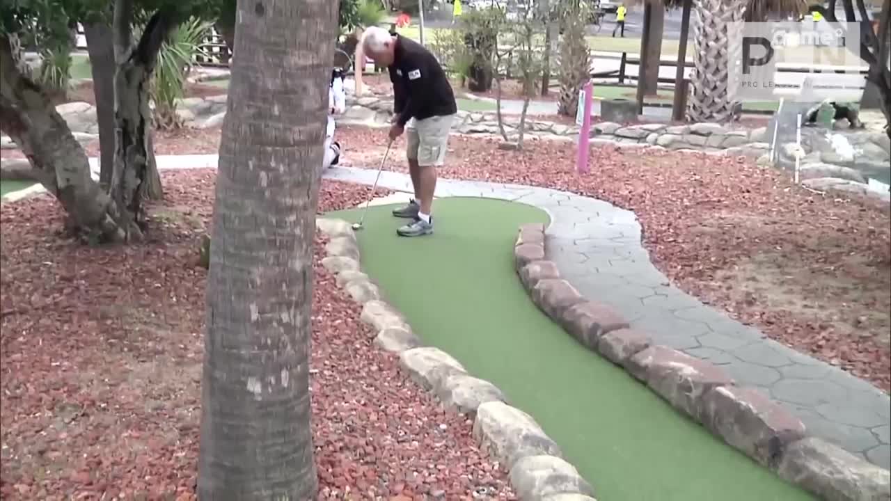 A man in shorts and a black shirt lines up a putt on a miniature golf course. The bright green turf curves around him, bordered by rough-hewn rocks.