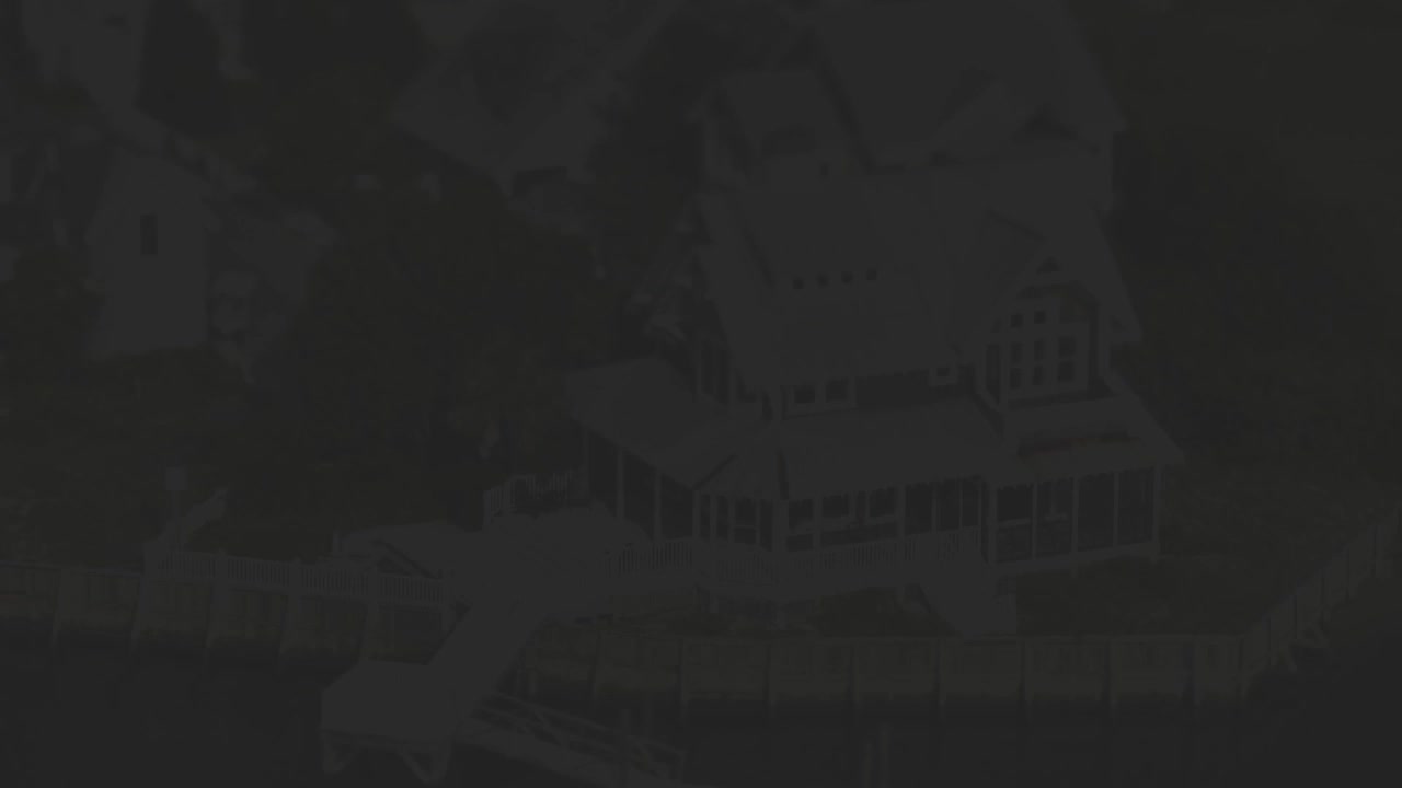 The drone hovers over a large, white house with a wrap-around porch.  A long, wooden dock extends from the property into dark water.