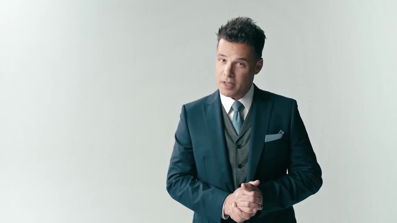 A man in a sharp suit stands with his hands clasped, speaking directly to the camera. He's presenting something important, perhaps a segment for Game+ in Canada.