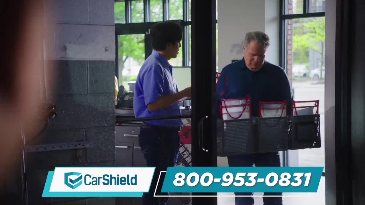 A man in a blue shirt is handing something to another man carrying a large, black caddy filled with items. They are standing near an open doorway.