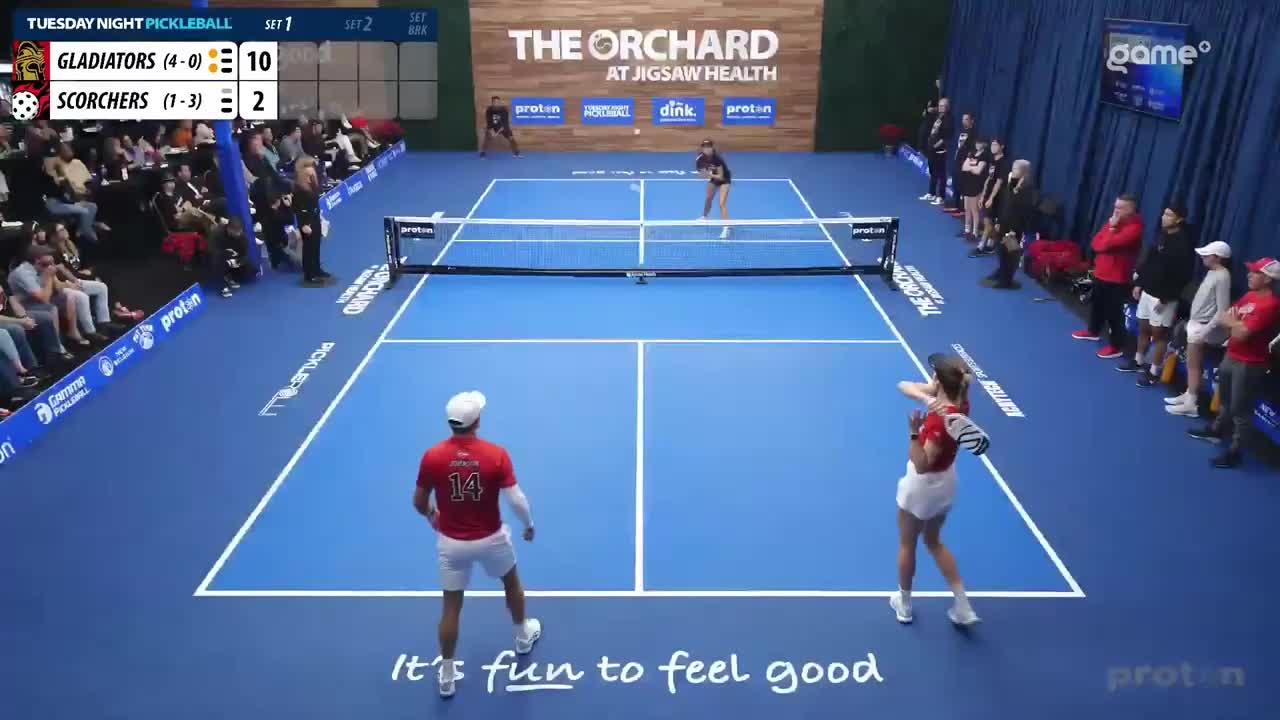 A woman in a red shirt and white shorts is about to hit a pickleball, while her male partner in a red shirt with the number 14 stands ready. The scoreboard shows the Gladiators leading the Scorchers 10-2 in the second set of Tuesday Night Pickleball.
