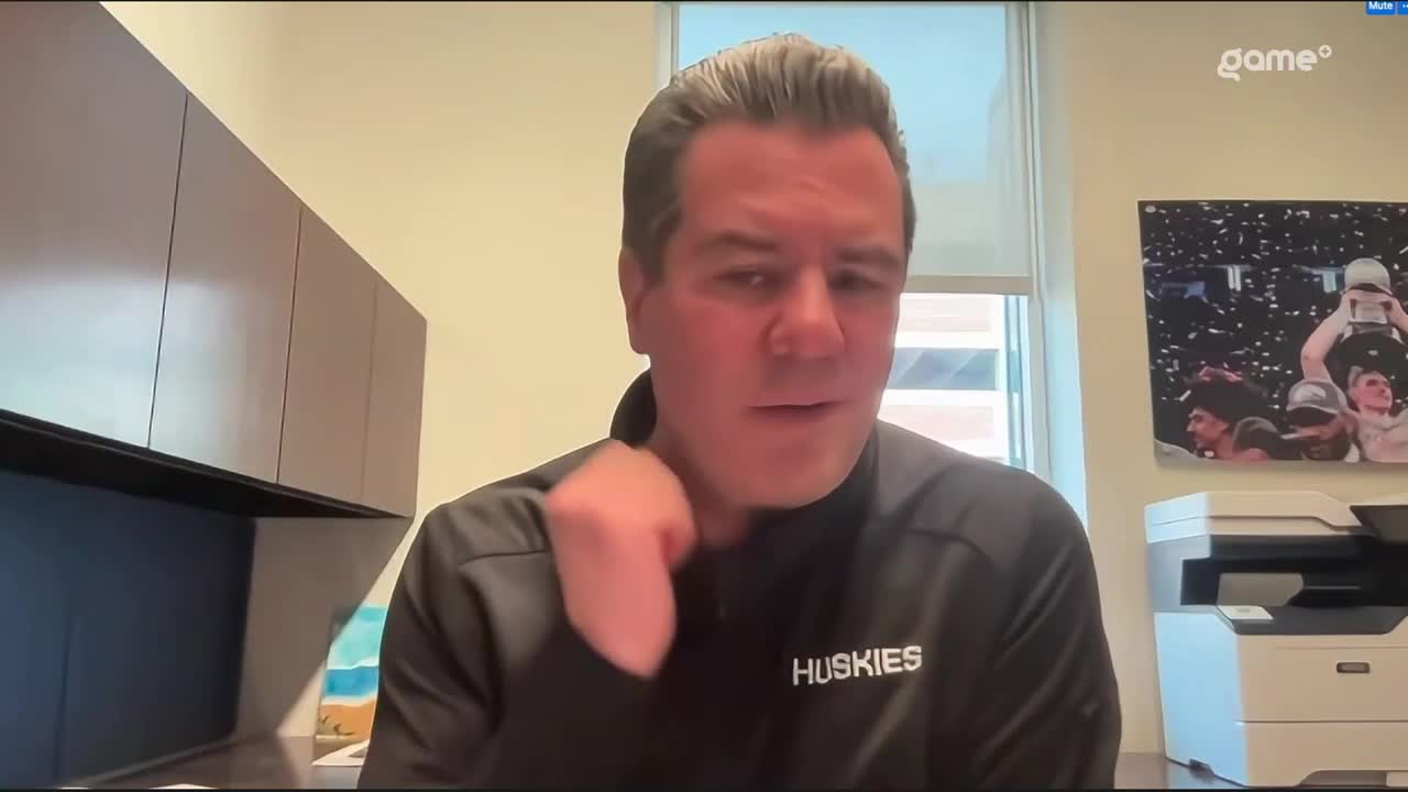 A man in a black jacket with "Huskies" on it speaks, his hand moving near his neck. Behind him, a Game+ logo appears above a photo of a championship celebration.