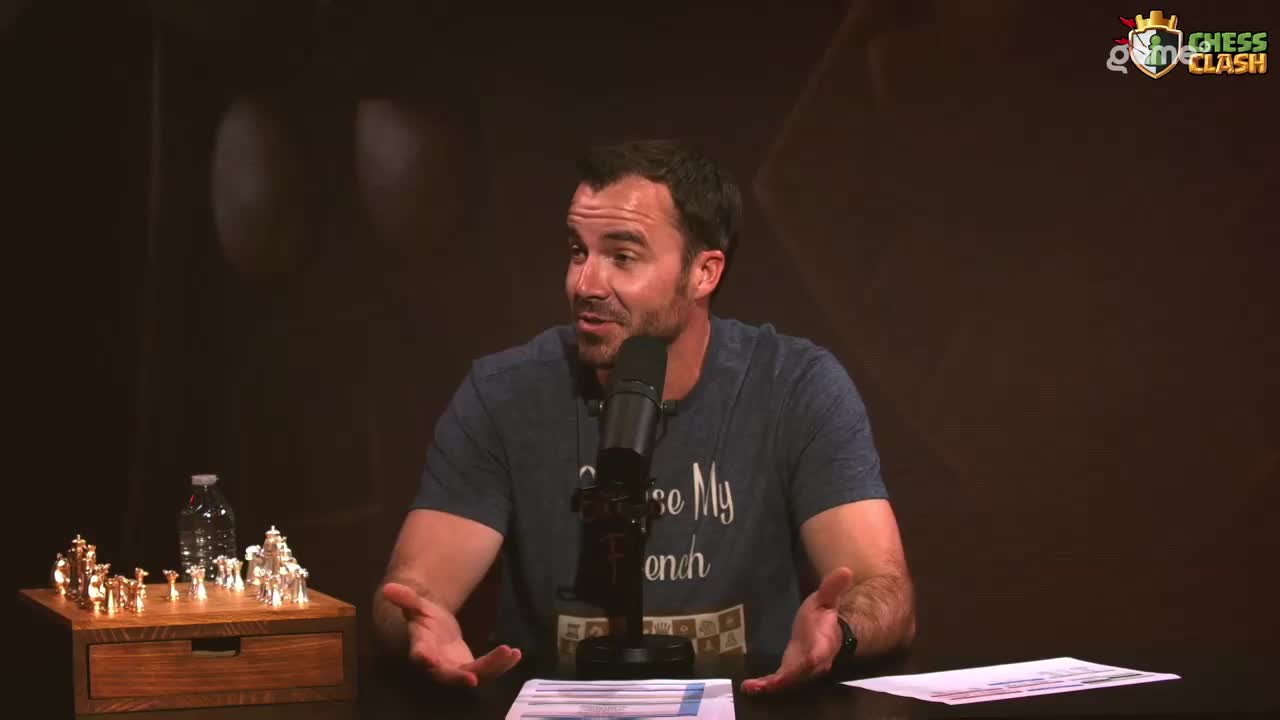 A man in a blue t-shirt gestures with his hands while speaking into a microphone. A small, ornate chess set sits on a wooden box to his left.