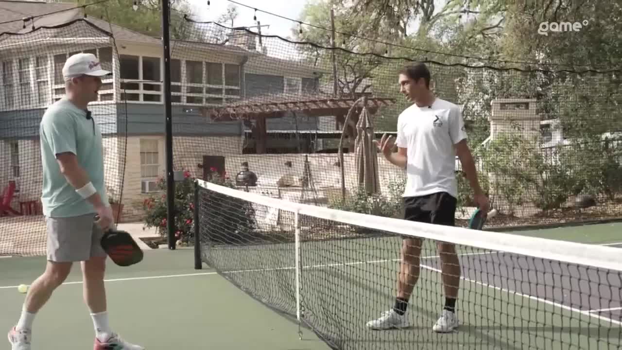 Two men stand on a pickleball court, one on each side of the net. The man on the left, wearing a light blue shirt and a white hat, holds a paddle. The man on the right, in a white t-shirt and black shorts, gestures with his hands.
