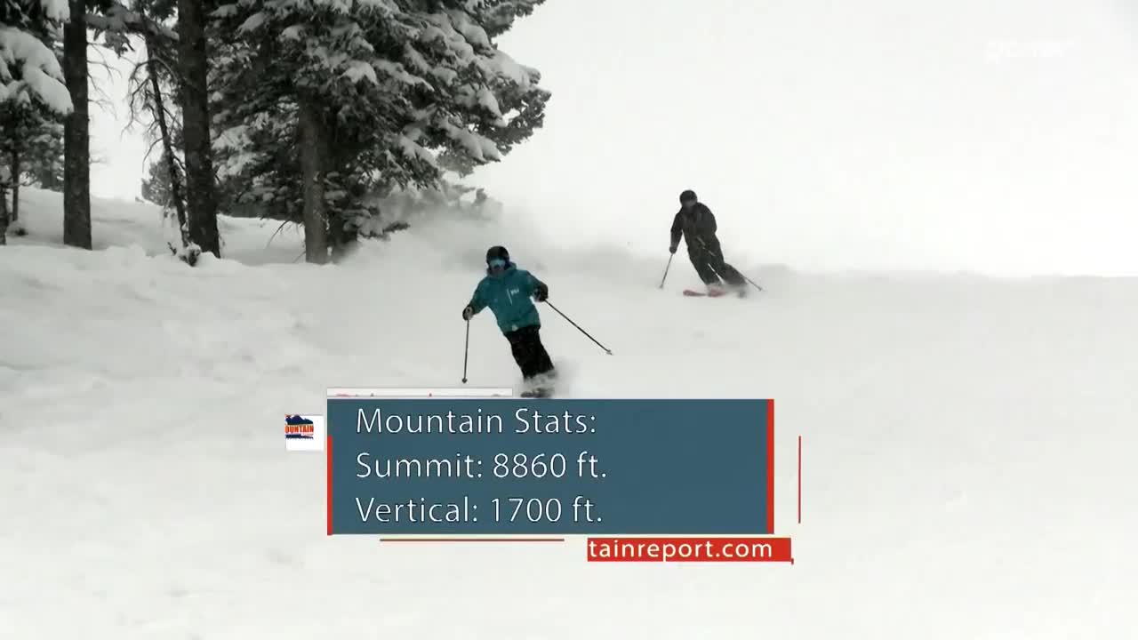 A skier in a teal jacket carves down the slope, sending up a spray of snow. Another skier follows behind, further up the hill.