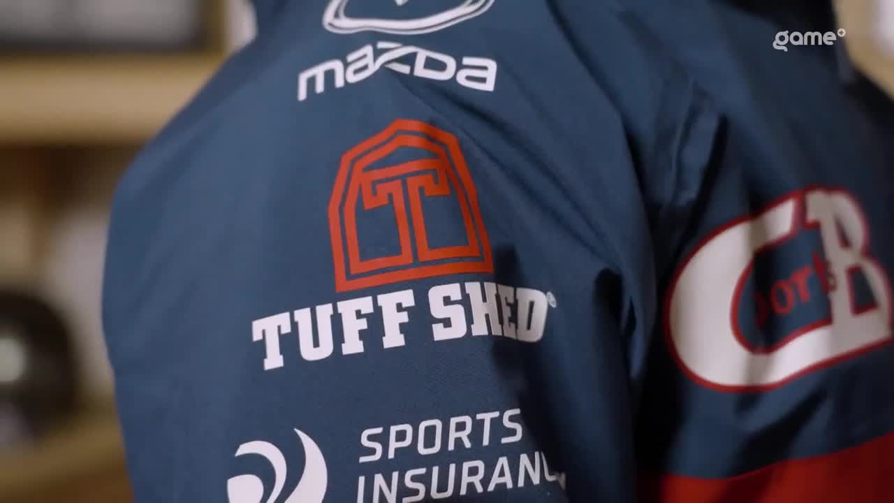 The back of a navy blue jacket displays the "Tuff Shed" logo in orange and white. A white "Game+" logo sits in the upper right corner.