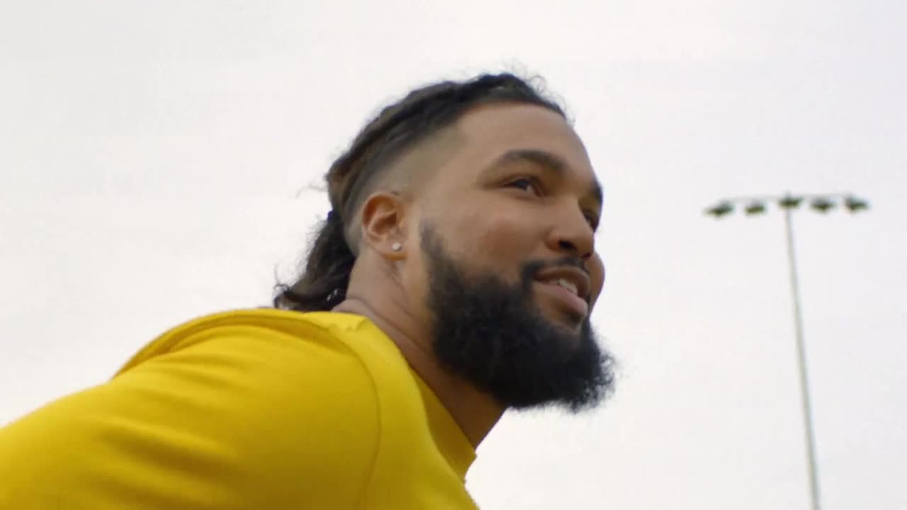 A man with braided hair and a beard, wearing a bright yellow shirt, turns his head to the side. In the distance, stadium lights stand against a pale sky.