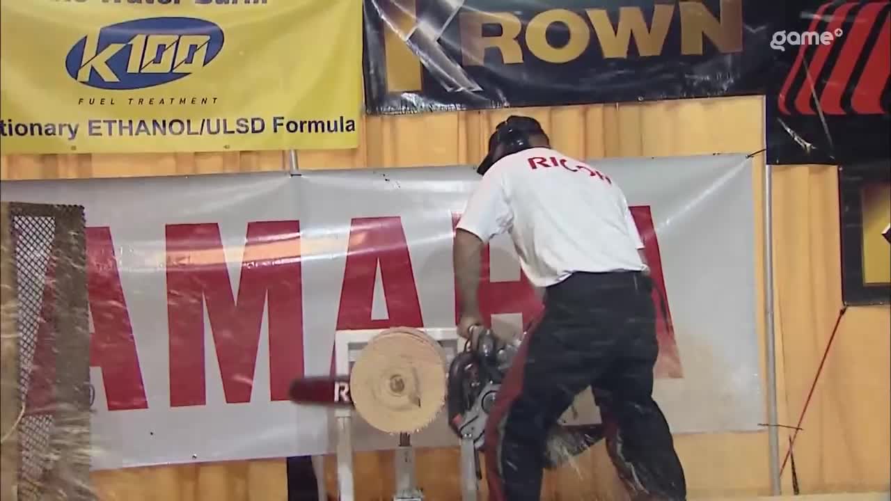 A lumberjack in a white "RICOH" shirt and black pants is powerfully swinging a chainsaw, sending wood chips flying. He's cutting through a thick log positioned on a stand, the blade a silver blur against the red "YAMAHA" banner behind him.