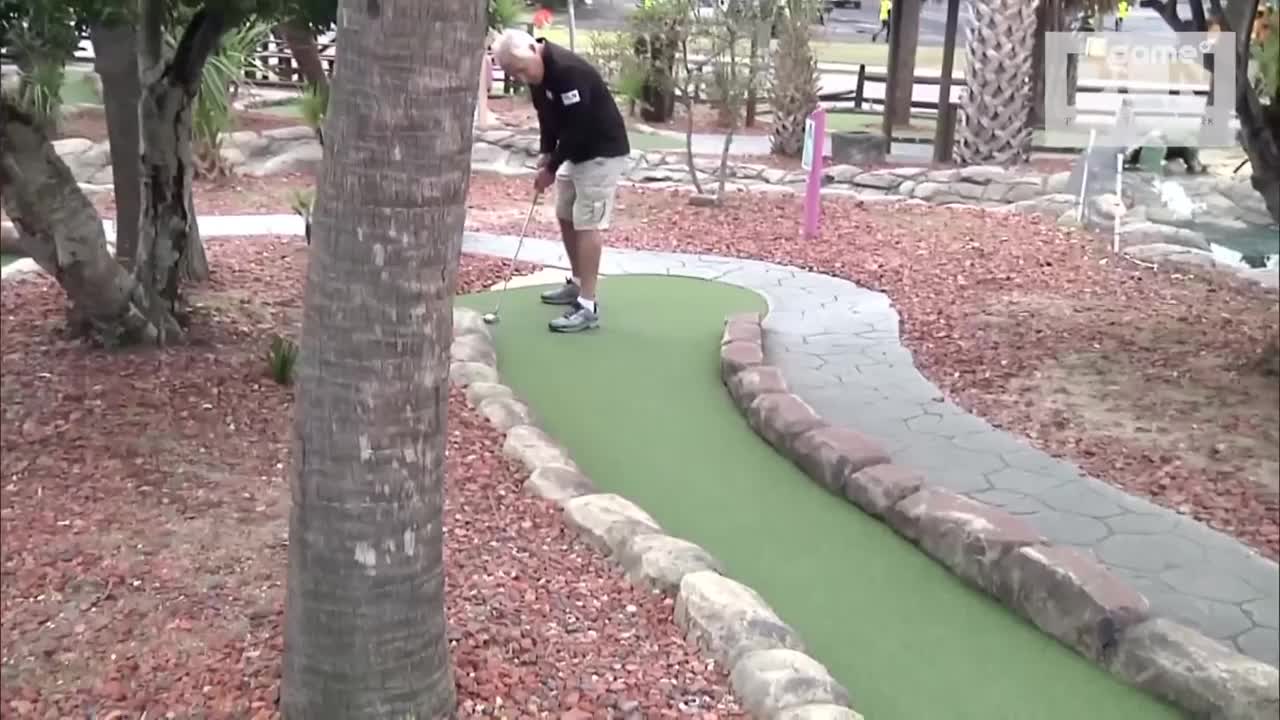 A man in a black shirt and khaki shorts lines up a putt on a miniature golf course. The ball rests on the bright green turf, just a few feet from the hole.