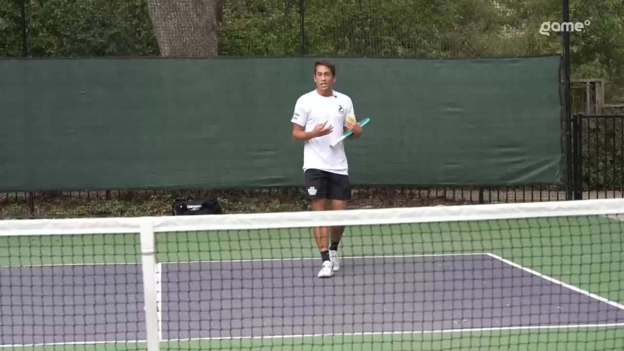 A tennis player in a white shirt and black shorts stands on the court, holding a racket and ball. He's mid-sentence, gesturing with his free hand as if explaining a point for Game+.